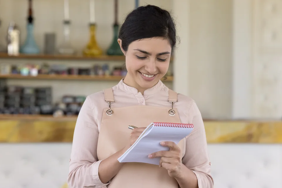A hospitality professional in an apron writing on a notepad in a cafe