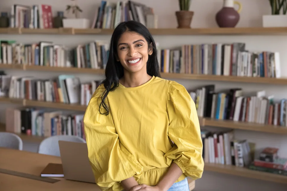 A smiling learner in a yellow blouse beside a laptop, with library bookshelves behind her